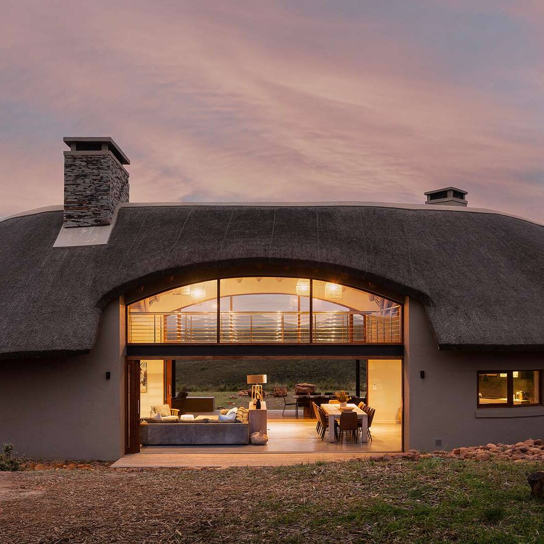 Exterior evening view of curved thatched form with wide opening to living pavilion, bushveld architecture