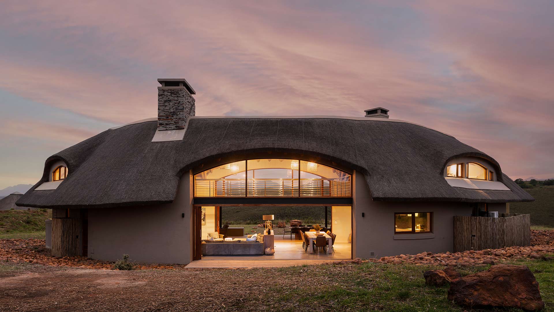 Exterior evening view of curved thatched form with wide opening to living pavilion, bushveld architecture